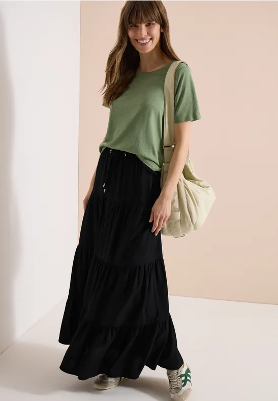 Femme en tee-shirt vert, jupe longue noire, sac clair et baskets.