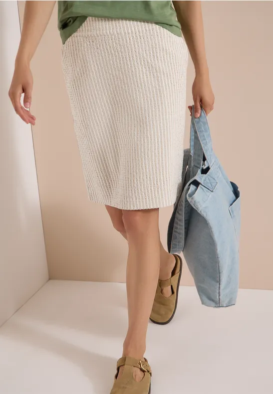 Femme en jupe crème côtelée, top vert, tenant une veste en jean bleue et portant des mocassins marron.