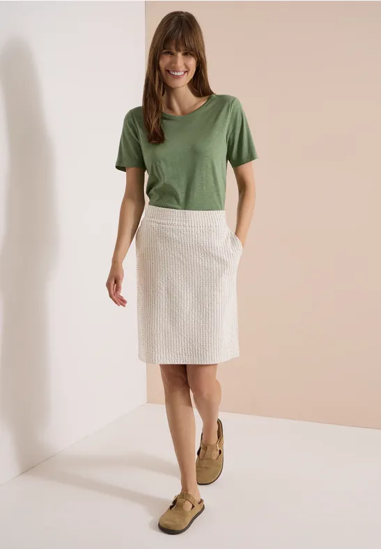 Femme en t-shirt vert et jupe blanche, chaussures marron, décor minimaliste.