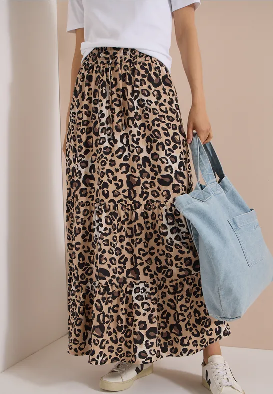 Femme porte une jupe longue léopard, t-shirt blanc, baskets blanches et un sac en jean.