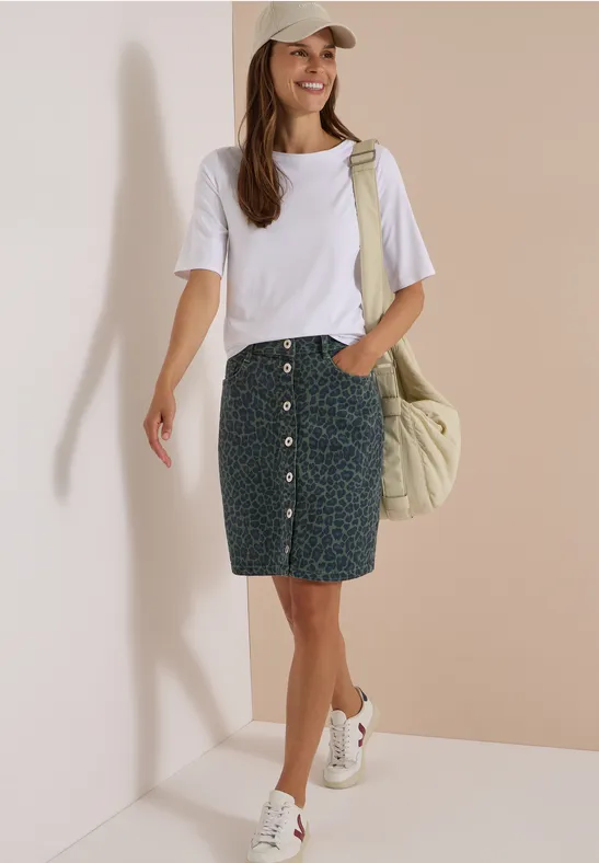 Femme en t-shirt blanc, jupe en jean à motifs et boutons, baskets blanches, casquette, sac épaule décontracté.