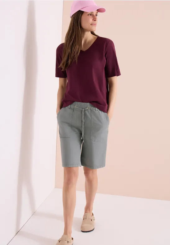 Person in V-Ausschnitt T-Shirt in Bordeaux, grauen Bermudas und beigen Sneakern, lehnt an weißer Wand.