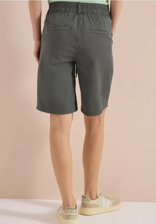 Person steht in grauen Shorts und hellen Sneakern, Rückenansicht vor beigem Hintergrund.