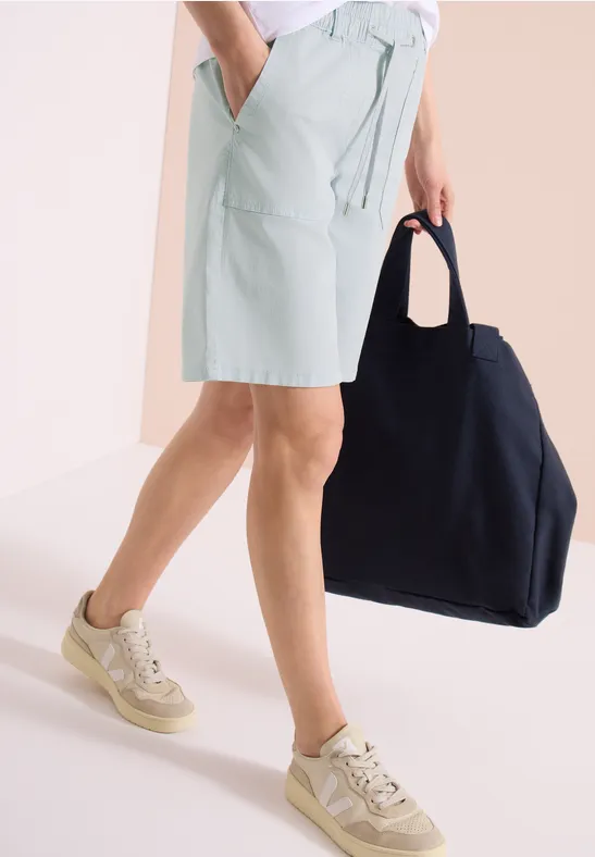 Eine Person trägt mintgrüne Shorts, beige Sneaker und hält eine große schwarze Tasche.