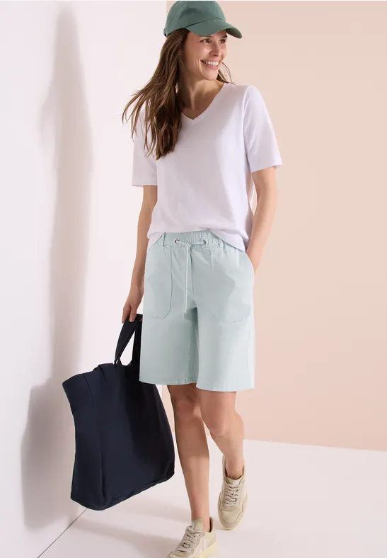 Frau in weißem T-Shirt, mintgrünen Bermudashorts und Cap trägt eine große Tasche.