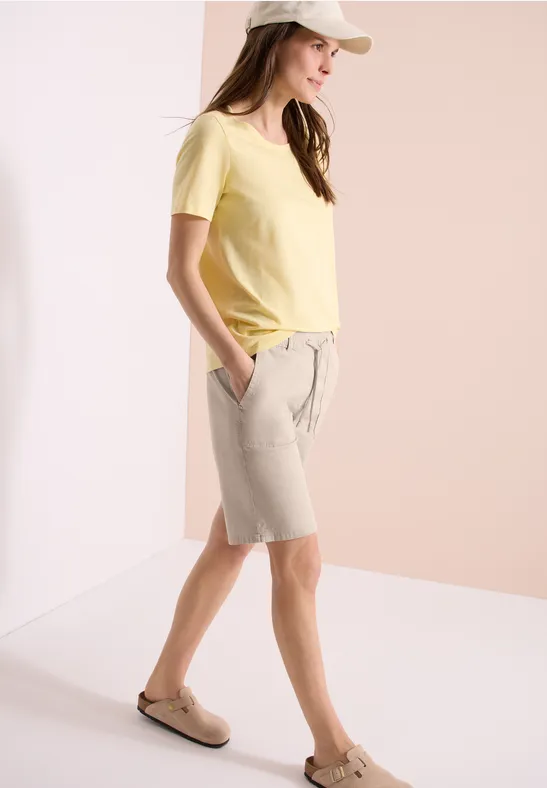 Frau trägt gelbes T-Shirt, beige Bermudas und Slipper vor hellem Hintergrund.