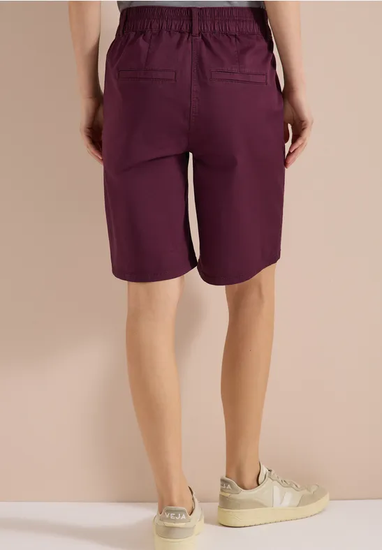 Rückenansicht: Person trägt violette knielange Shorts mit Taschen und helle Sneakers vor neutralem Hintergrund.