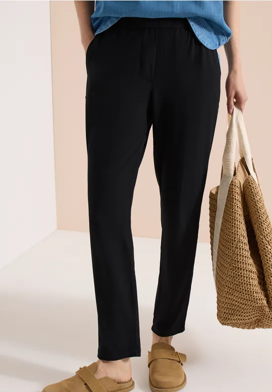 Person trägt schwarze, lässige Hose und Hausschuhe, hält eine geflochtene Tasche.