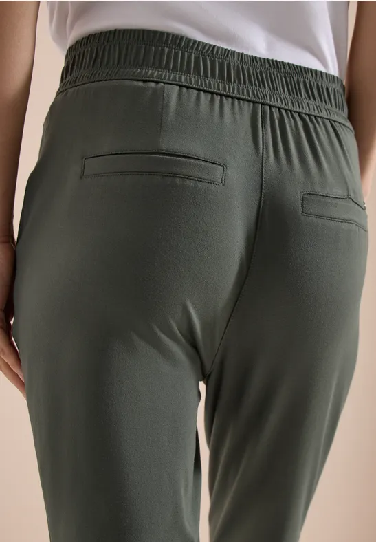 Rückenansicht einer Person in dunkelgrüner, elastischer Hose mit Bund und zwei Gesäßtaschen, kombiniert mit weißem Shirt.