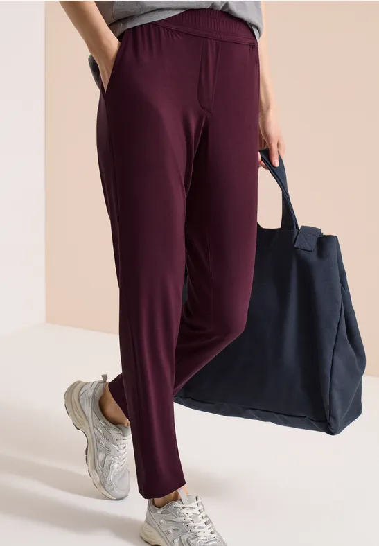 Person trägt lässige beerenfarbene Hose mit weißen Sneakern und hält eine große blaue Tasche.