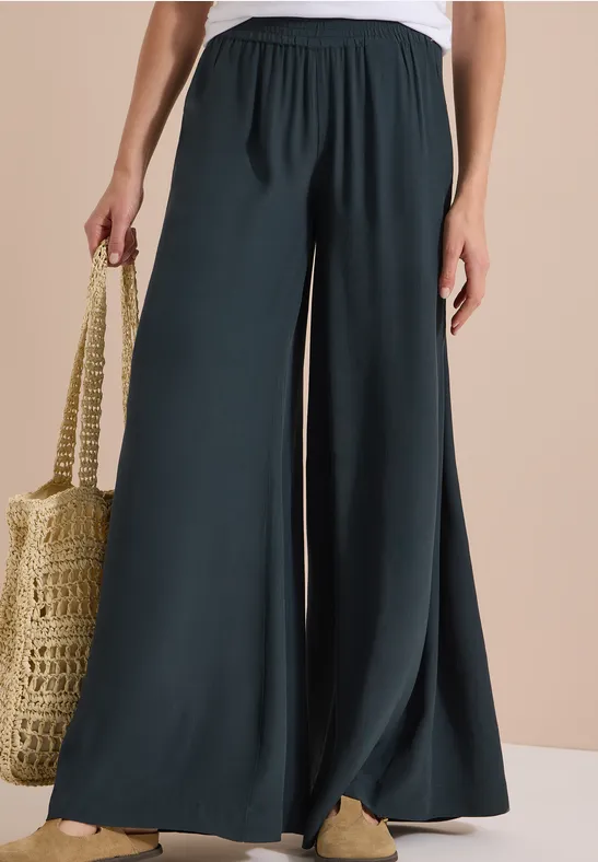 Pantalon ample vert foncé taille haute porté avec un sac en osier et des chaussures beiges.