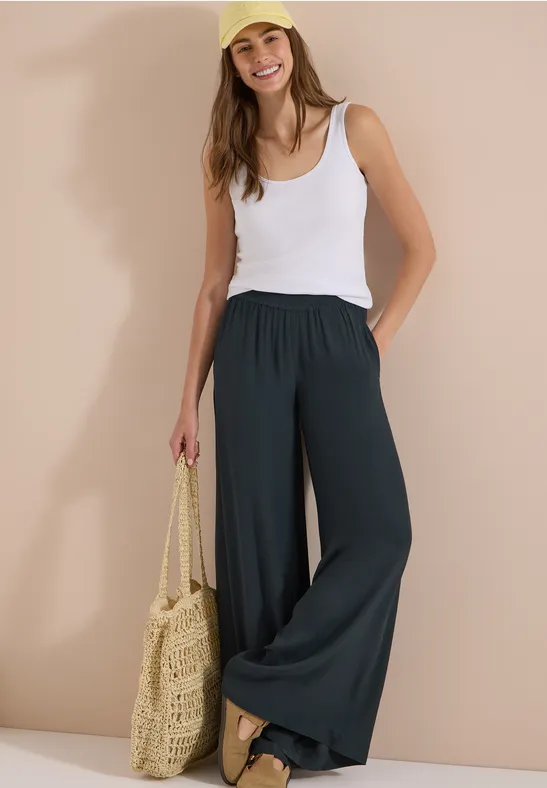 Femme avec débardeur blanc, pantalon large noir et sac en paille pour un look d'été décontracté.