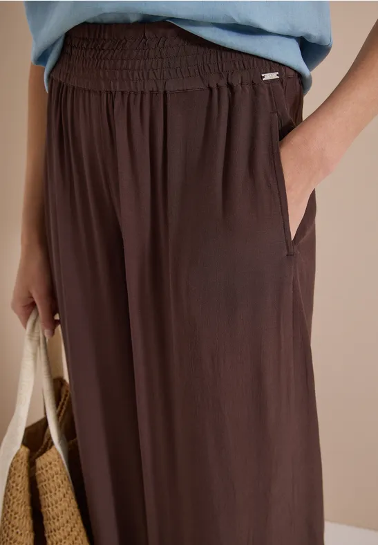 Gros plan d'un pantalon marron fluide à taille élastique et poches latérales, main dans la poche, sac en paille.