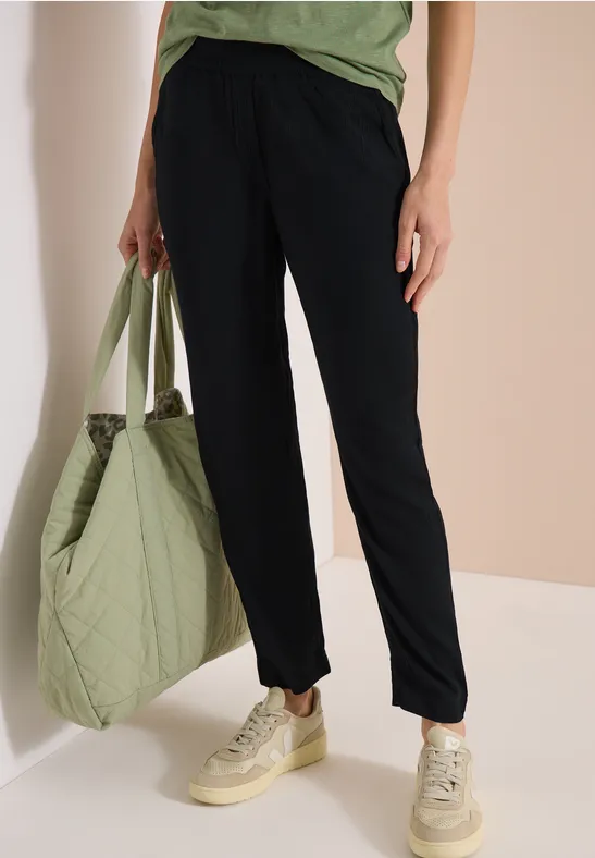 Person trägt dunkelblaue Hose, hellgrünes Shirt, hält einen gesteppten Shopper und beige Sneaker.