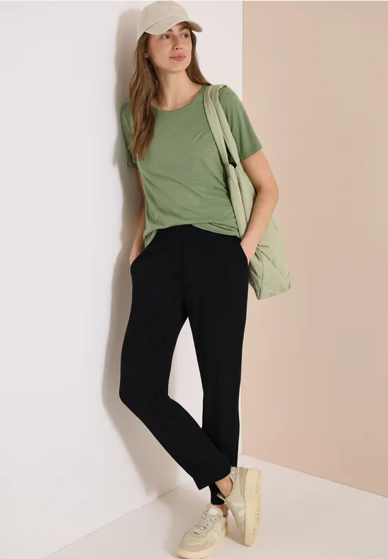 Eine Frau lehnt an einer Wand, trägt ein grünes T-Shirt, schwarze Hose, weiße Schuhe und eine grüne Tasche.