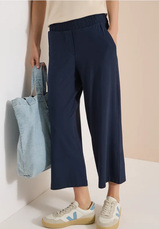 Person trägt dunkelblaue Culotte-Hose mit elastischem Bund, weiße Sneaker und hält eine hellblaue Stofftasche.