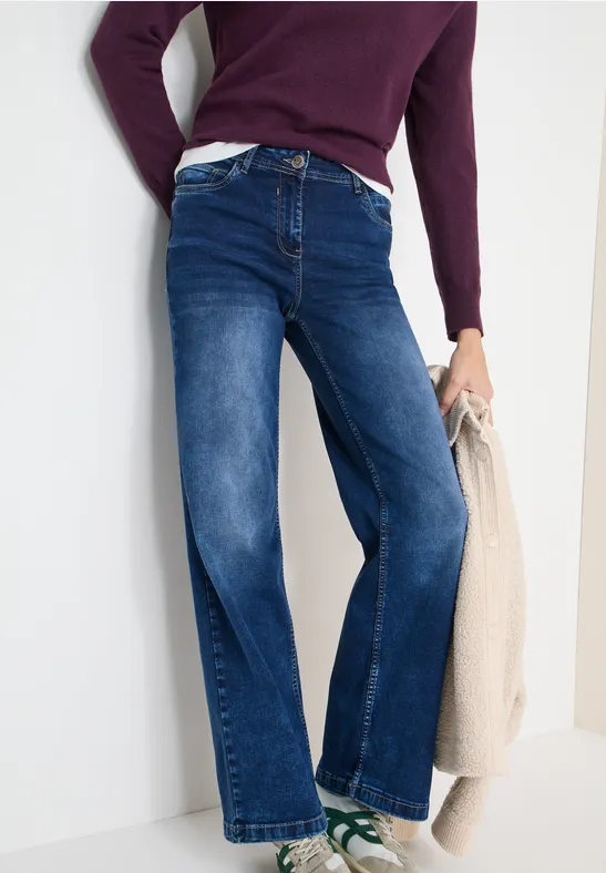 Person trägt weite, dunkelblaue Jeans, lila Pullover, weiße Sneaker und hält eine beige Tasche.