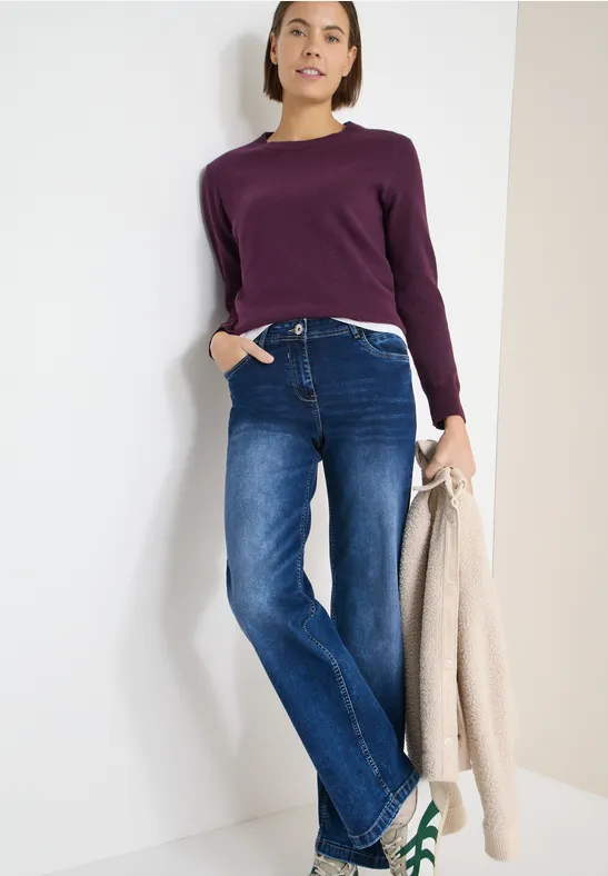 Frau in lila Longsleeve, weiter blauer Jeans, trägt helle Jacke in der Hand, lässige Pose vor hellem Hintergrund.
