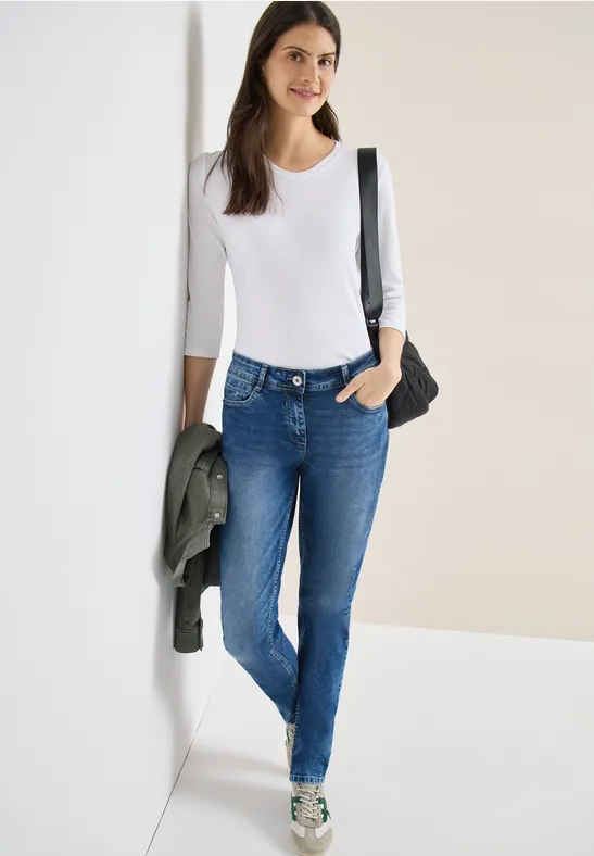 Frau in weißem Langarmshirt, blauen Jeans, grauen Sneakers und schwarzer Umhängetasche lehnt an Wand.