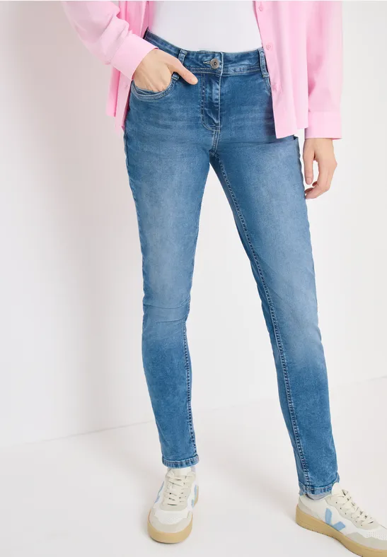 Een vrouw draagt strakke blauwe jeans, een roze vest en witte sneakers.