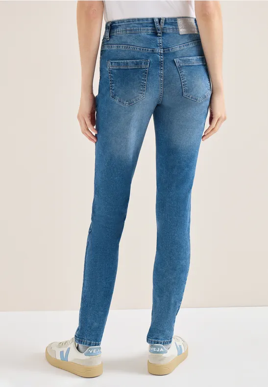 Achteraanzicht van een persoon in blauwe slim-fit jeans en witte sneakers tegen een neutrale achtergrond.