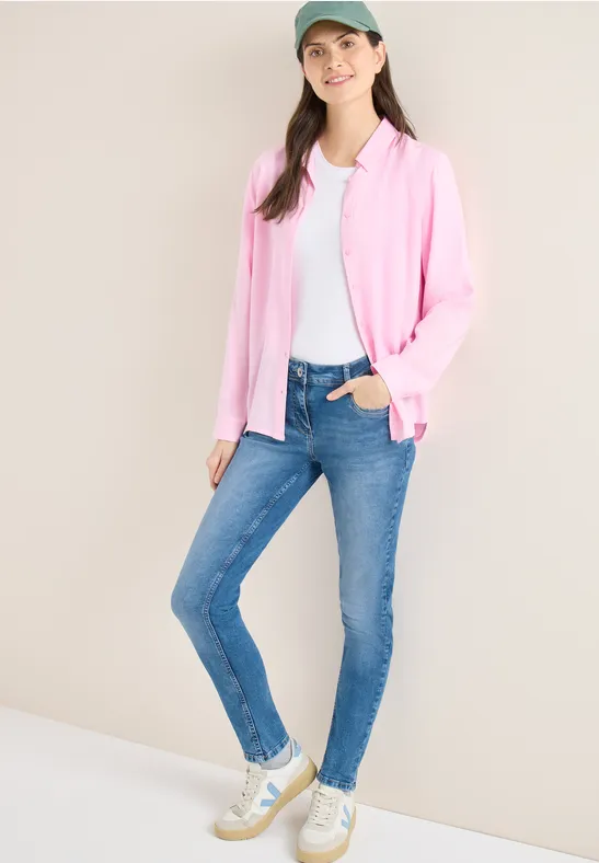 Vrouw in roze vest, wit shirt, blauwe jeans en witte sneakers.