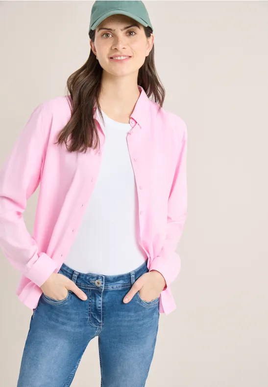 Persoon met een roze vest, wit topje en blauwe jeans tegen een lichte achtergrond.