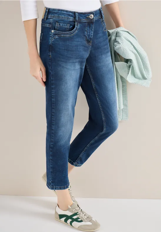 Femme en jean 7/8 bleu, baskets blanches à accents verts, tenant un gilet vert menthe.