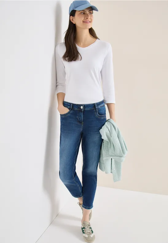 Femme en t-shirt blanc, jean bleu et mocassins argentés, appuyée contre un mur avec une veste menthe.