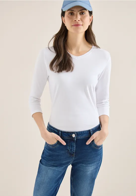 Femme en t-shirt blanc aux manches 3/4, jean bleu et casquette, mains dans les poches.
