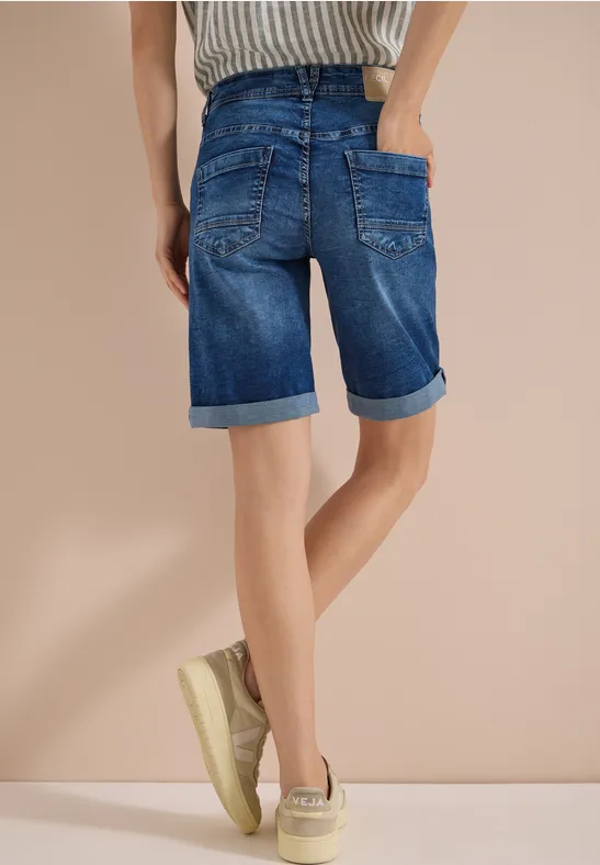 Vue arrière d'une personne portant un short en jean retroussé aux genoux et des baskets beiges sur fond neutre.