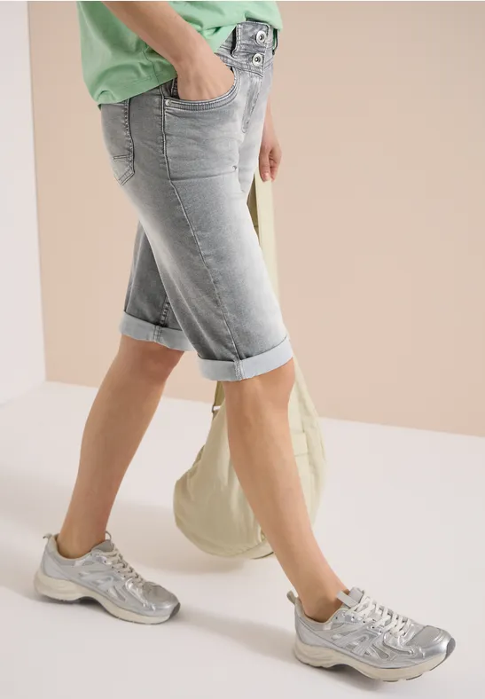 Beine in hellblauen Bermudashorts, silbernen Sneakern, grünem Oberteil und beiger Canvas-Tasche.