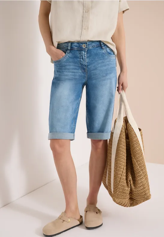 Person in hellblauen knielangen Jeans-Shorts, beiger Bluse, beigen Slippern und Strohtasche.