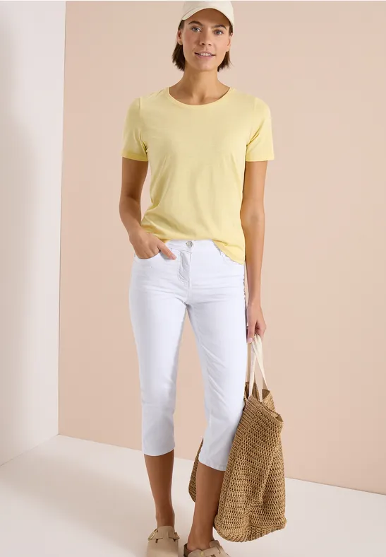 Frau in gelbem T-Shirt, weißen Caprihosen und geflochtener Tragetasche.