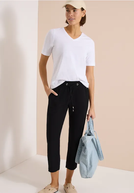 Frau in weißem V-Neck-T-Shirt, schwarzen Jogginghosen und weißen Sneakern, mit Jeans-Tasche in der Hand.