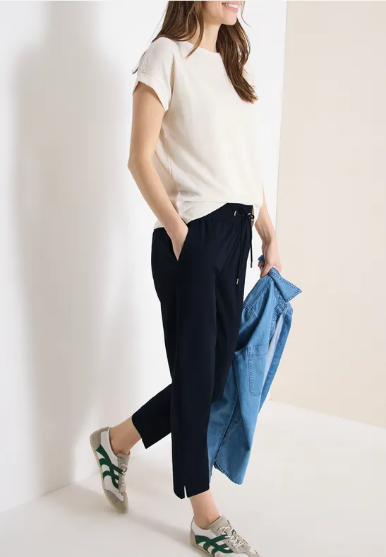 Frau in weißem T-Shirt, dunkelblauer Hose und grünen Sneakern, hält blaue Jeansjacke.