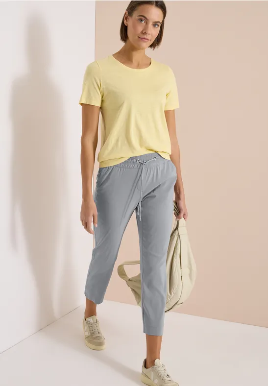 Frau in gelbem T-Shirt, grauen 7/8-Hosen und Sneakern steht lässig und hält eine helle Tasche.