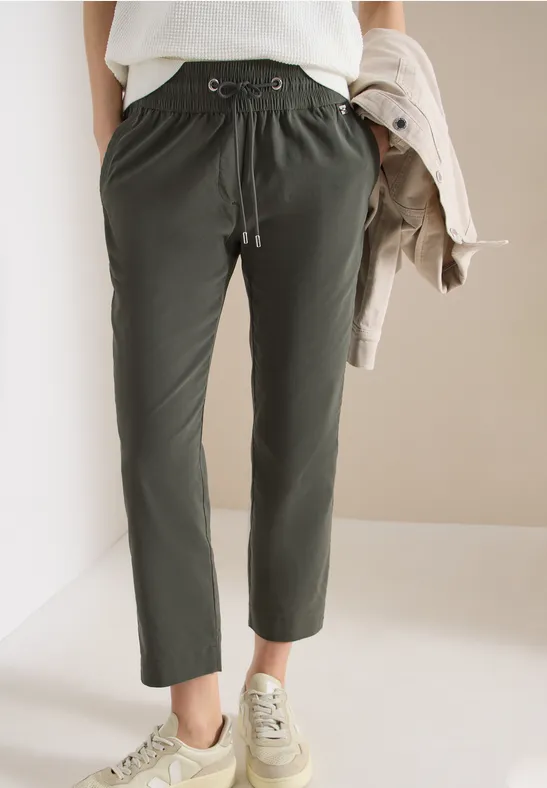Personne portant un pantalon vert foncé à cordon et des baskets beiges, tenant une veste beige.