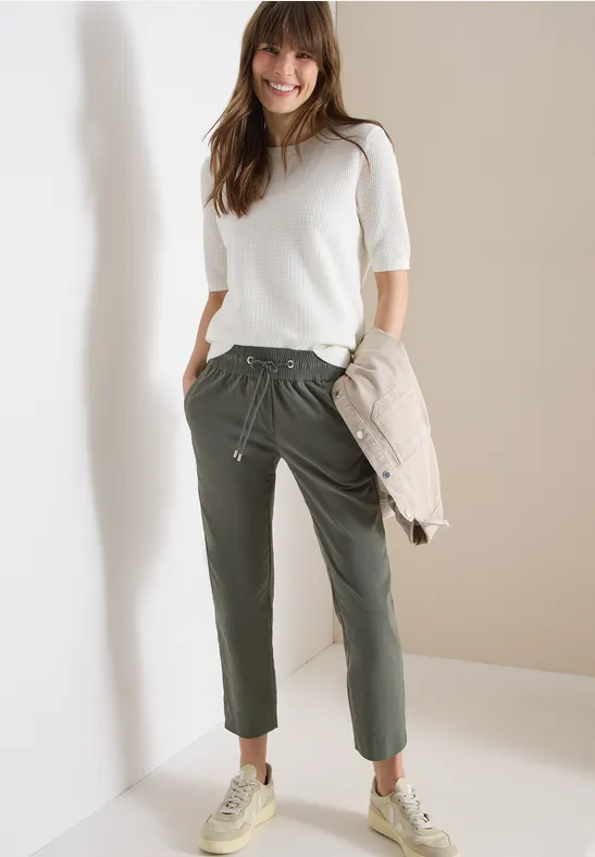 Femme en t-shirt blanc, pantalon vert olive et baskets beiges appuyée contre un mur, tient une veste