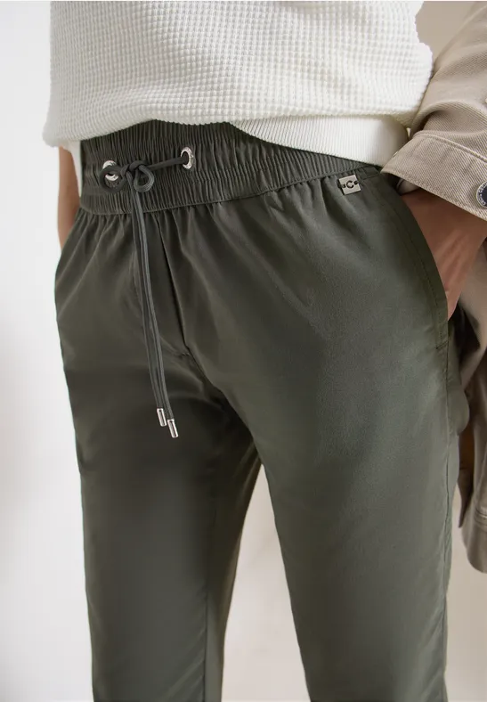 Un pantalon de jogging vert olive avec cordon, porté avec un haut clair et une veste décontractée sur le bras.