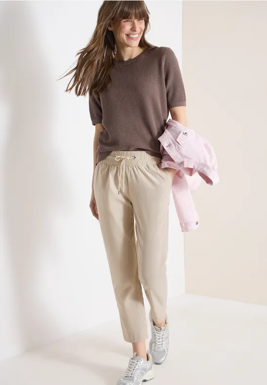 Frau in braunem Shirt, beigen Hosen und Sneakern hält eine rosa Jacke.