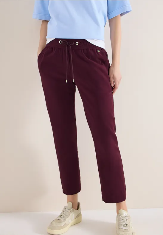 Person trägt hellblaues T-Shirt, bordeauxrote Hose mit Kordelzug, Hände in den Taschen und weiße Sneaker.