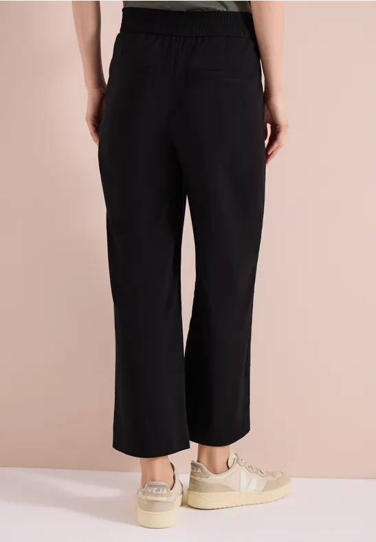 Frau von hinten in schwarzen, weiten, knöchellangen Hosen mit elastischem Bund und hellbeigen Sneakern.