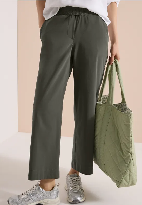 Eine Person trägt olivgrüne, weite Hose mit elastischem Bund, silberne Sneaker und hält eine gesteppte grüne Tasche.