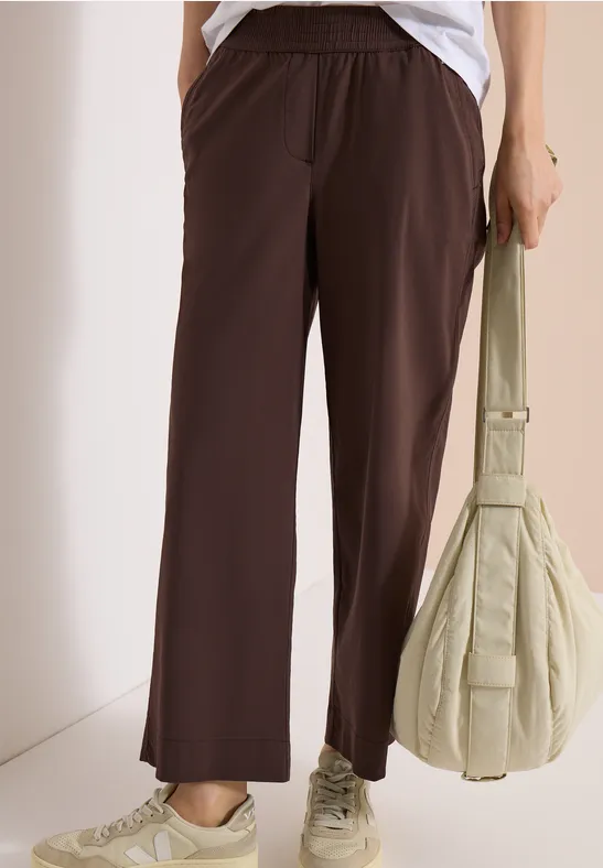 Person trägt weite braune Hose, weiße Sneakers und hält eine helle, gepolsterte Handtasche.