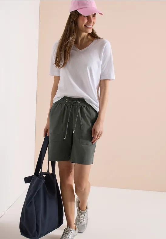 Junge Frau in weißem T-Shirt, khakigrünen Shorts, rosa Cap und Sneakers mit dunkelblauer Tasche.