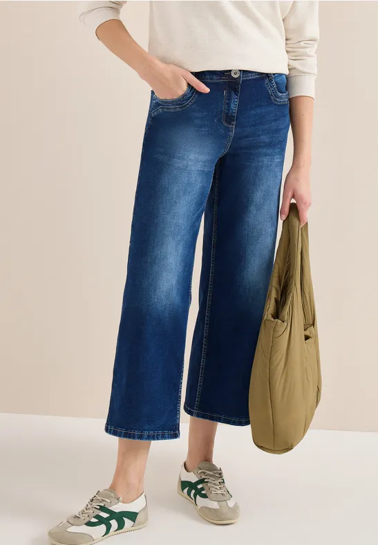 Person in weißem Oberteil, weiten, knöchellangen Jeans, weißen Sneakern mit grünen Details und grüner Tasche.