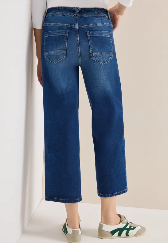 Rückansicht einer Person in kastig geschnittenen, knöchellangen blauen Jeans und weißen Sneakern mit grünen Streifen.