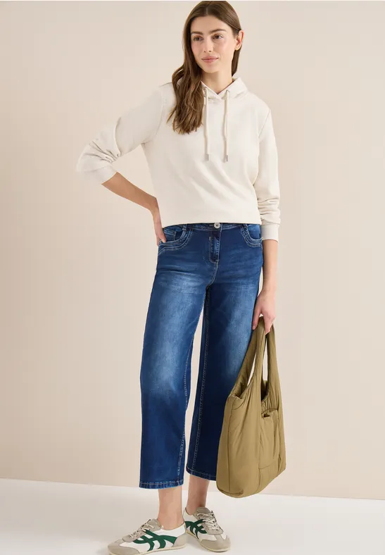 Person trägt weißen Hoodie, weite blaue Jeans, grüne Sneaker und eine olivgrüne Tasche.