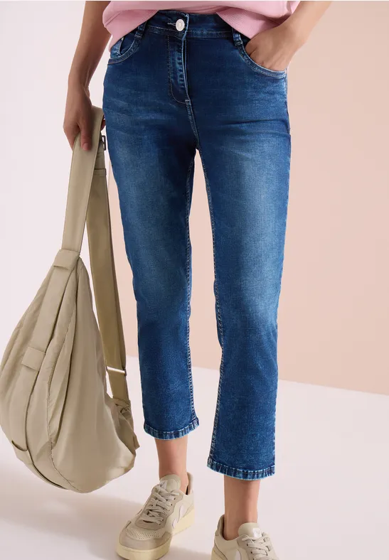 Frau trägt knöchellange, blaue Jeans, rosa Oberteil, beige Sneakers und eine bequeme beiger Tasche.